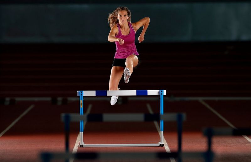 A female athlete in a purple vest leaping over a hurdle.