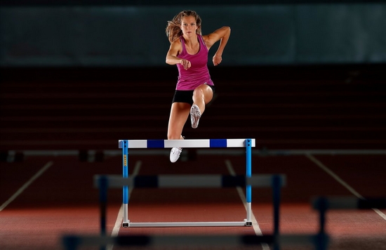 A female athlete in a purple vest leaping over a hurdle.