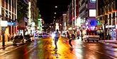 Two people skateboard down a city street at night, colourful lights from buildings reflecting in the wet road surface.