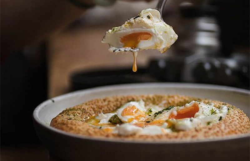 A bowl of food shows eggs cooked with spinach in a bread bowl; a spoonful of egg is lifted.