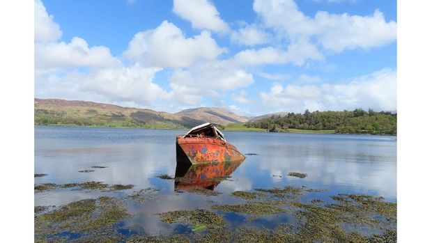 A rusty red boat sits in the middle of a lake on a sunny day
