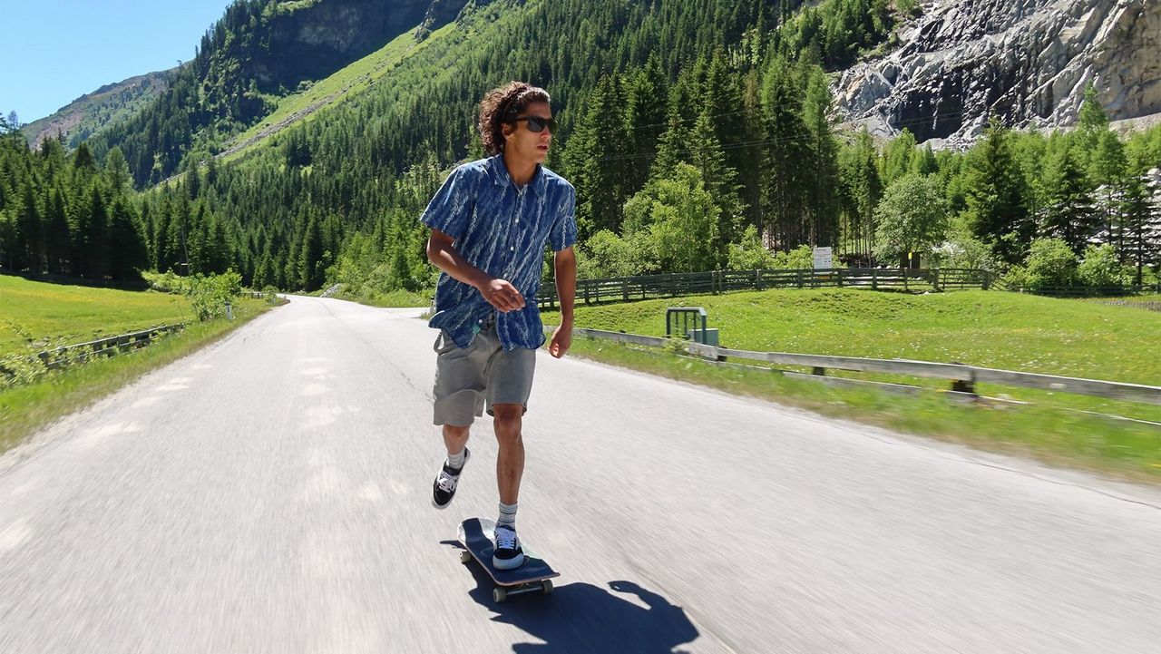 A man skateboards down a road past mountains covered in trees.