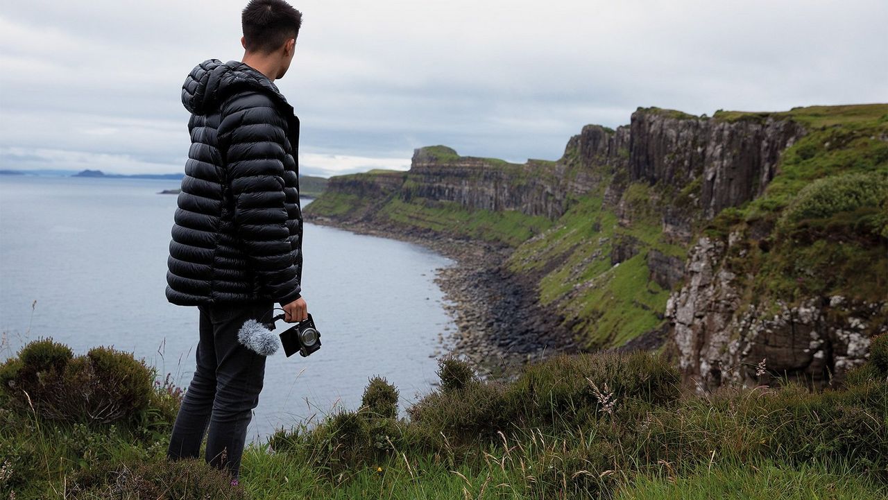 Mike Gray stands with his back to the camera looking at the Isle of Skye coastline, holding a Canon PowerShot G7 X Mark III with mic attached.