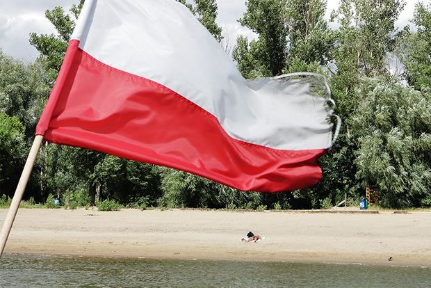 The Polish flag flutters in front of a beach, deserted but for one figure on the sand, with lush woods behind.