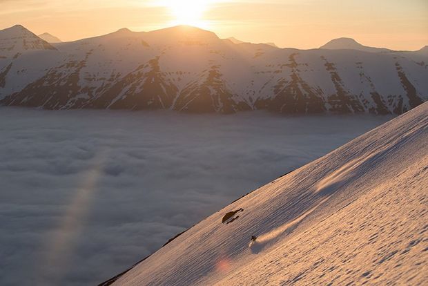 A wide shot shows a mountainous landscape with someone skiing down, low sun behind.