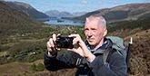 Outdoor photographer Alan Rowan stands on top of a hill in front of a lake and fields, holding a compact Canon camera.