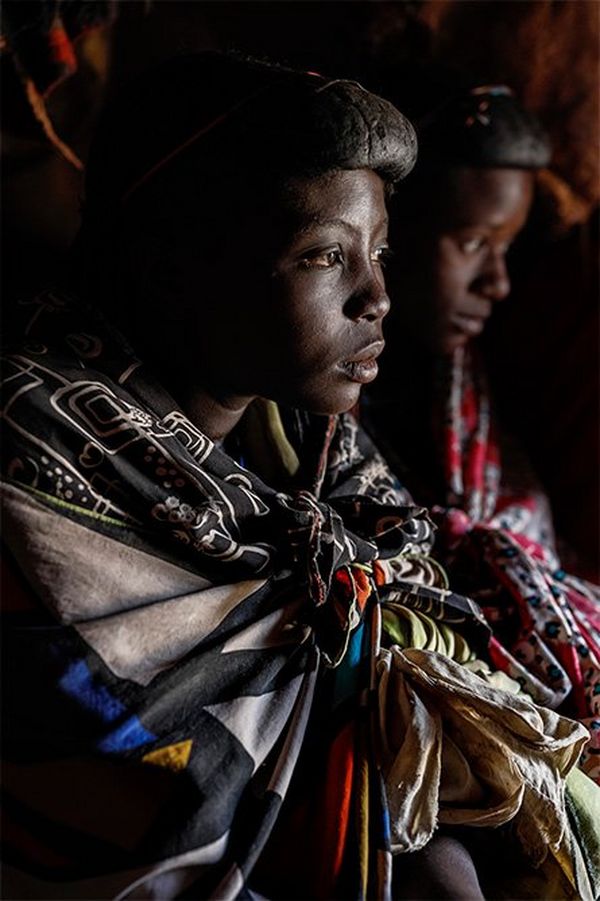 A thoughtful young Himba woman wearing four or five differently-patterned scarves, all knotted at the front, with a companion out-of-focus in the background.