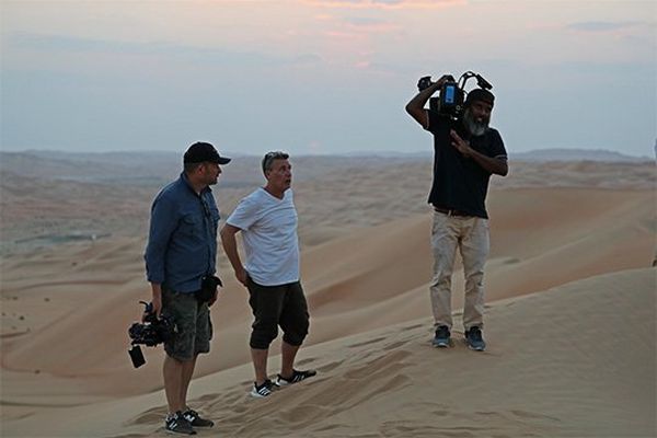 Three men in a desert, two holding Canon video cameras ready to film.