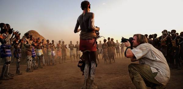 A photojournalist crouches beside a man in tribal African dress to take his photograph, while others dressed in traditional tribal clothes clap and dance.