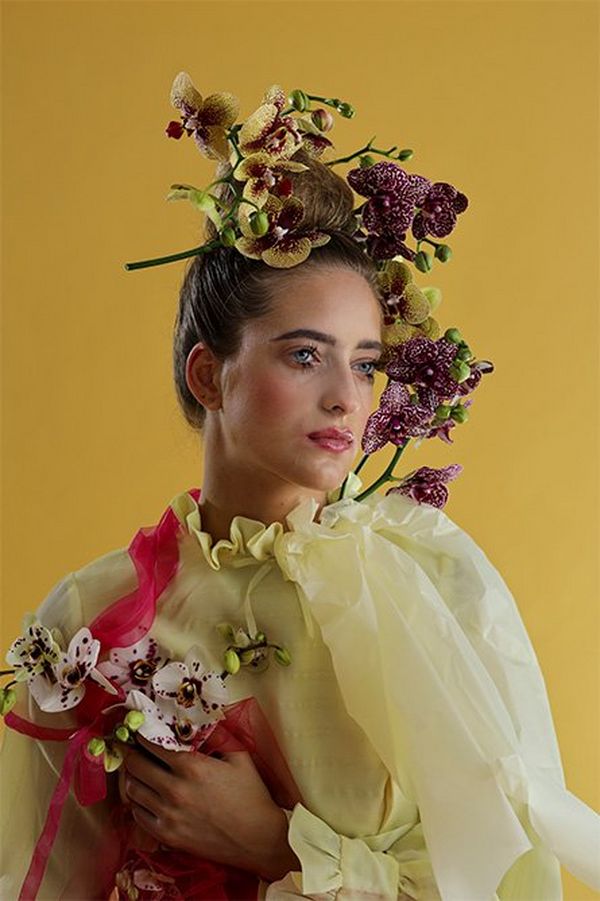 A white model has orchids and fabric draped over and around her, against an ochre backdrop in a studio portrait with a yellow theme. 