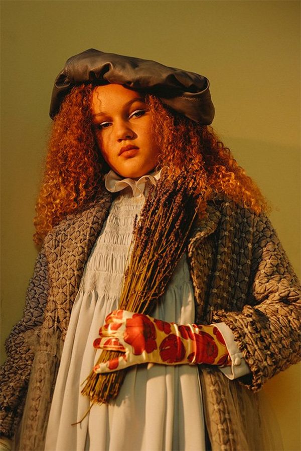 A woman with tightly curled red hair, wearing a rustic-looking smock, coat, hat and red flowery glove, looking slightly down at us, holding a sheaf of dried lavender.