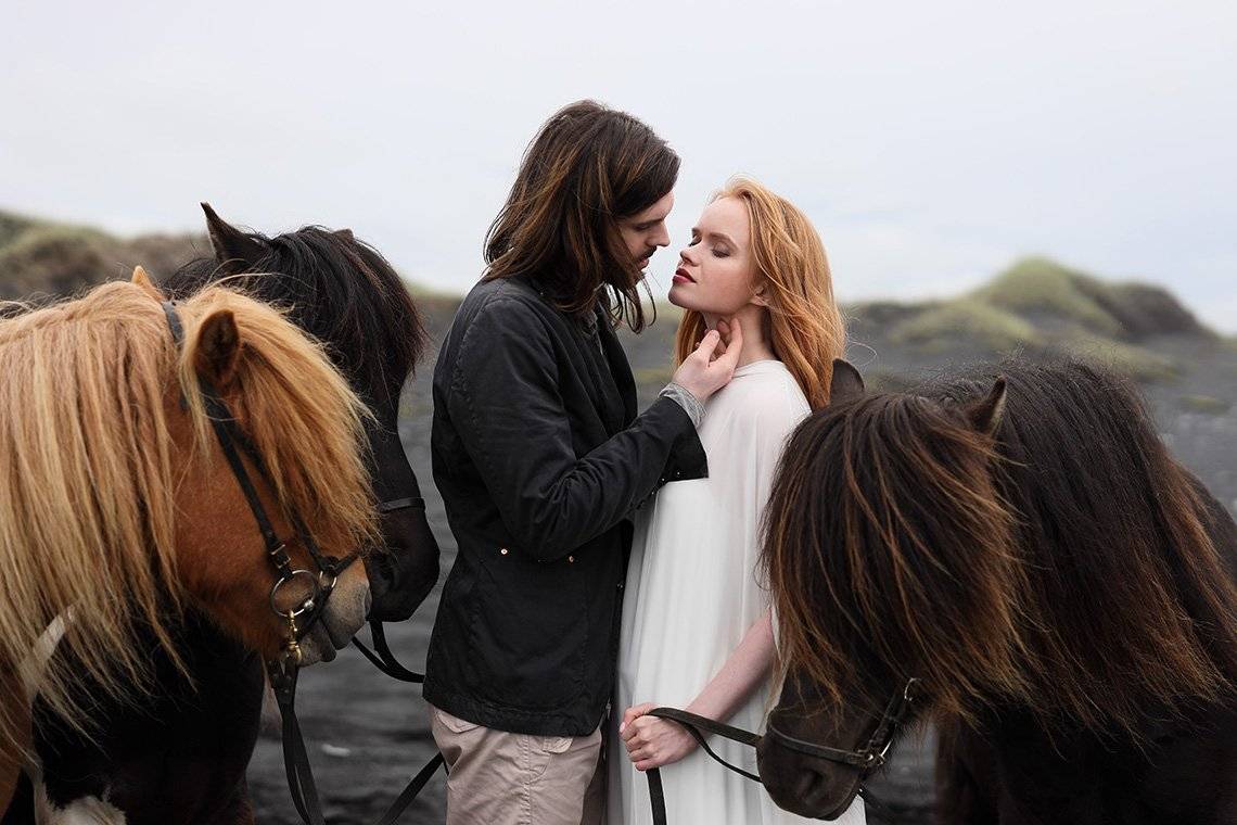A man with long dark hair caresses a strawberry blonde woman in a white dress, among a group of ponies in a natural landscape.