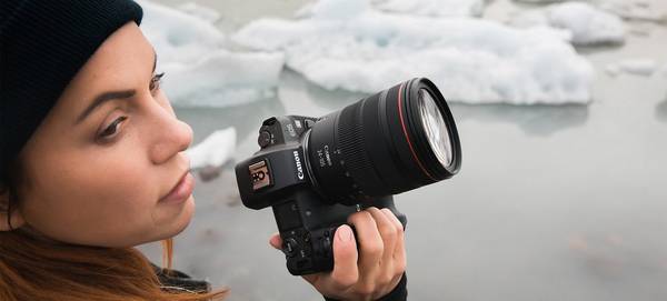 Photographer Katya Mukhina stands in front of a body of water with icebergs in it, holding a Canon EOS R in her right hand.