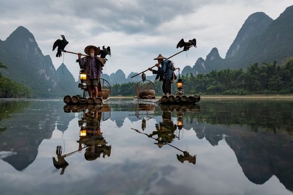 Two fishermen stand on their boats, each with a staff over his shoulder. A cormorant perches at the end of one staff and both ends of the other. The entire scene is reflected in the still water.