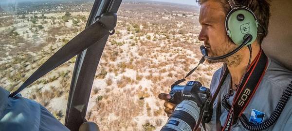 Neil Aldridge in a light aircraft looking out over the African grasslands below, holding a Canon EOS 5D camera.