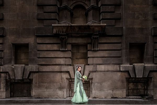 A bride in an intricately detailed mint green dress stands in front of the stone facade of a London building. Taken by wedding photographer Sanjay Jogia.