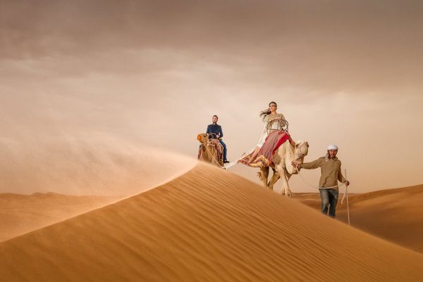 A couple riding camels are led by a guide over sand dunes in Dubai. Taken by wedding photographer Sanjay Jogia.