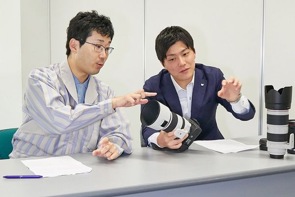 Mr Okuda points to the Canon RF 70-200mm F2.8L IS USM lens held by Mr Kawai as they sit at a table.