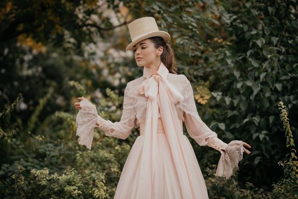 A model in a cream-coloured wedding dress and top hat poses in front of a blurred background of greenery.