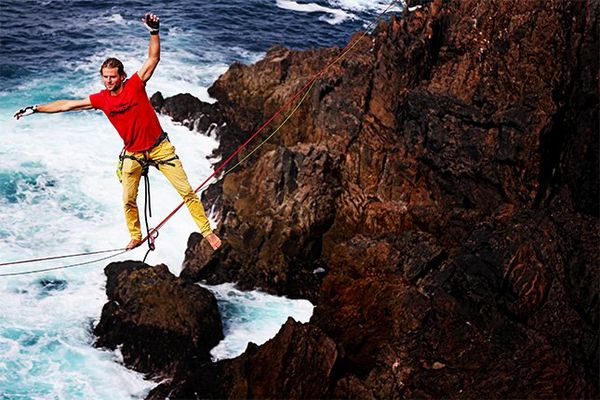 A man catches his balance on one foot, arms waving, standing on a rope suspended over the sea with the rocky shore visible below.