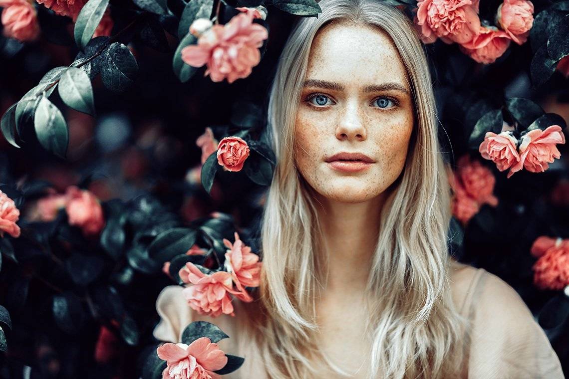 A blonde woman is surrounded by pink flowers and dark green leaves. Photo by Rosie Hardy with a Canon RF 85mm F1.2L USM lens.