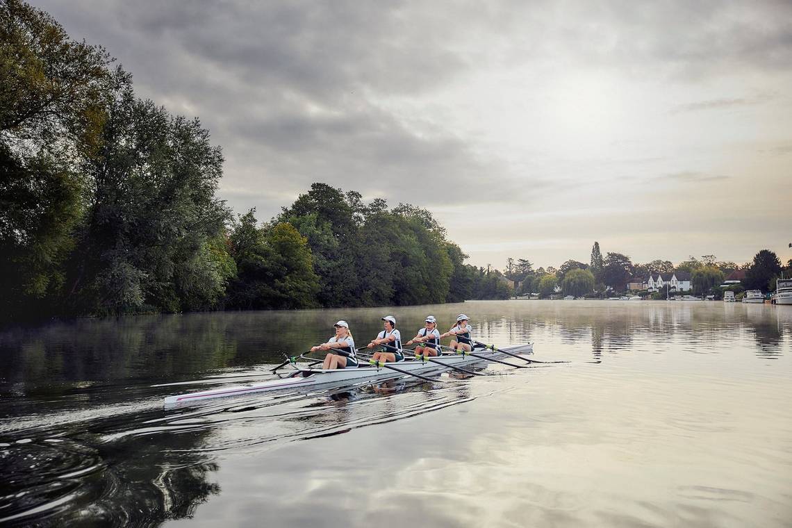 Four women row a narrow boat along a wide river at dawn.