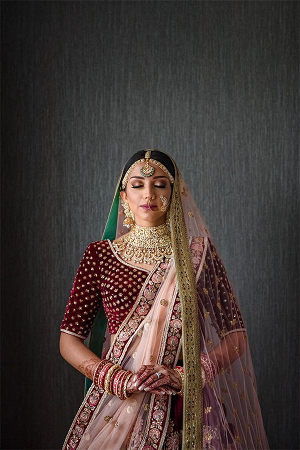 An Indian woman in a deep red wedding sari embroidered with gold, plus gold jewellery and henna patterns on her hands, stands peacefully in front of a dark grey wall.