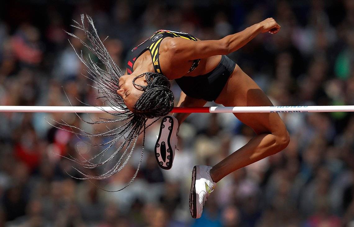 A female athlete soars over the high jump, her braided hair swirling around her head.