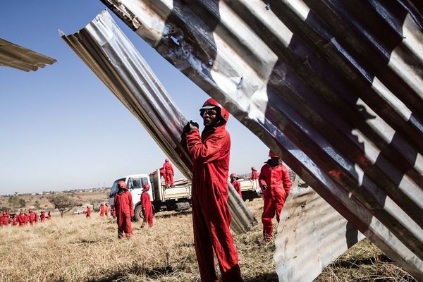 Construction workers wearing red boiler suits hold up sheets of corrugated metal in a field. 