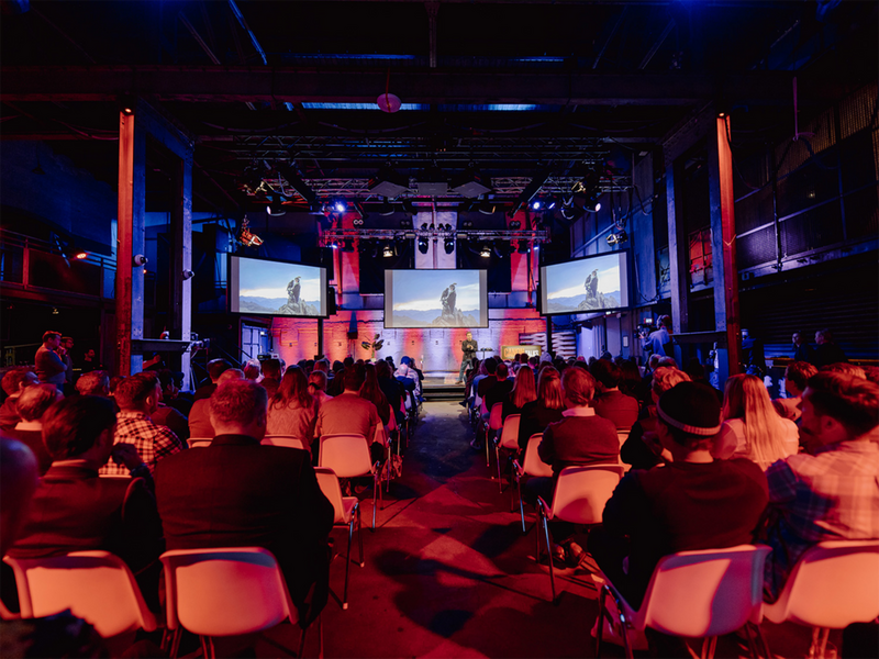 Sitting audience at an event, picture taken from the back with 3 screens and a scene shown in the distance.