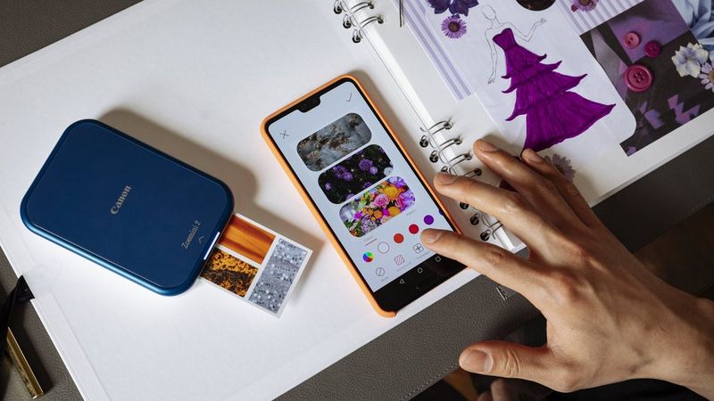 Navy blue Canon Zoemini 2 printing a picture on ZINK paper sticker with a smartphone showing Mini Print app with the printed picture on the screen. 