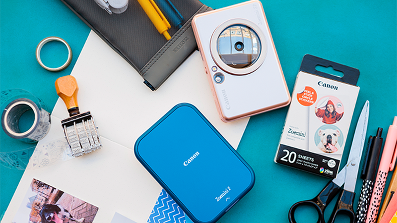 Canon Zoemini 2 in navy blue and Canon Zoemini S2 in white on a teal surface surrounded by tape, stamps and paper packaging.
