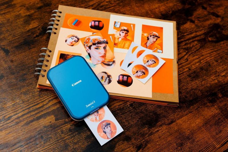 A Canon Zoemini 2 sits on top of a scrapbook covered in small photographs, printing two circular stickers of selfies.