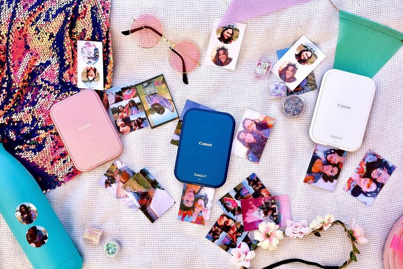 Three Canon Zoemini 2 models lie amidst printed photos, sunglasses, hairbands and a water bottle on a colourful mat. 