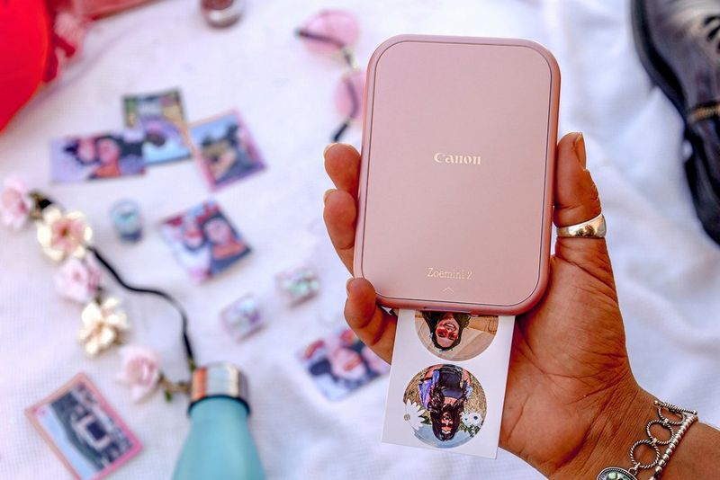 A hand holding a Canon Zoemini 2 compact photo printer with two circular photo stickers emerging from it.