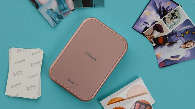Rose gold Canon Zoemini 2 on a blue surface surrounded by printed stickers and ZINK paper.