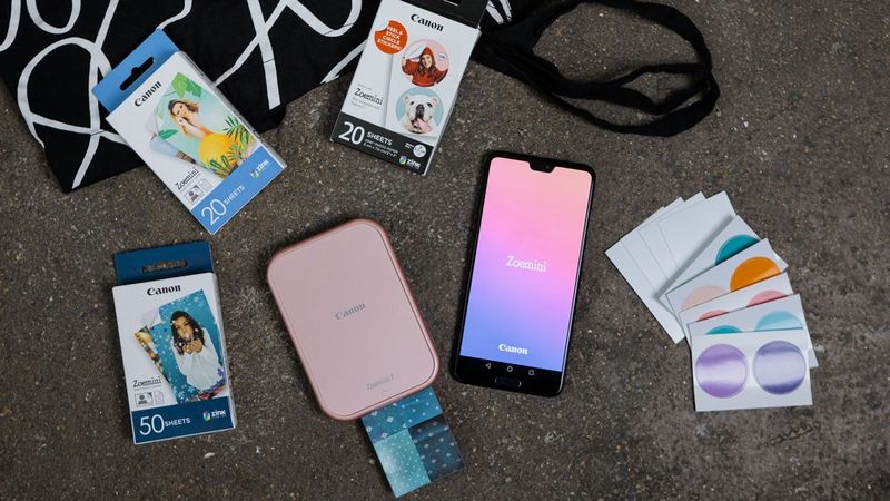 Rose gold Canon Zoemini 2 printing an image, surrounded by ZINK paper packages and printed stickers, with a smartphone with Mini Print app on it.