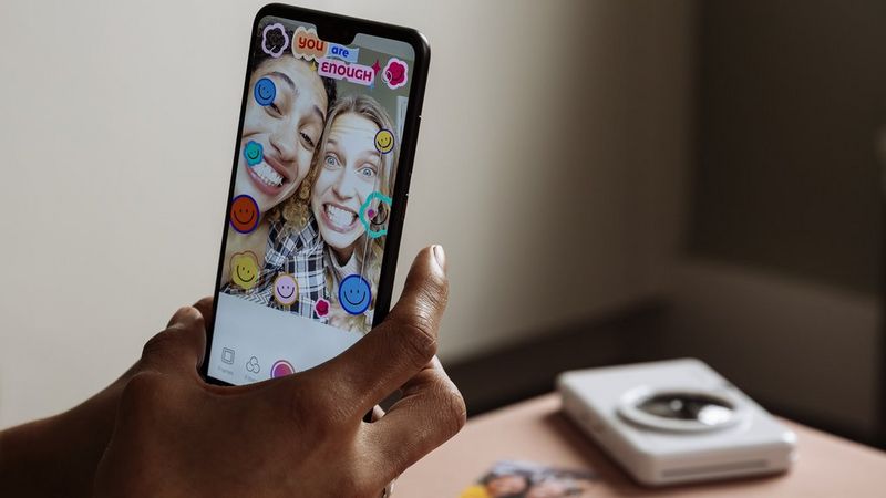 A woman holding the smartphone with Mini Print app showing a picture of two women with stamps and frames applied. Pearl white Canon Zoemini S2 is seen in the background. 