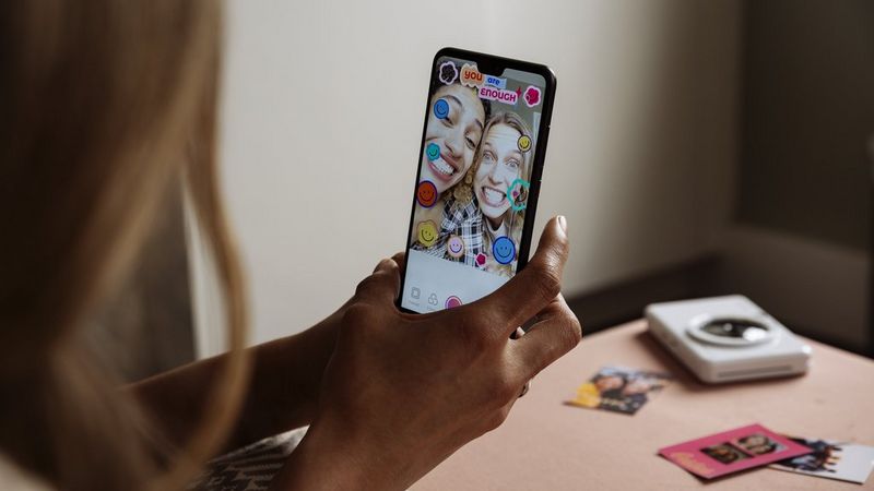 A woman holding a smartphone showing Mini Print app with a picture of two friends and stamps applied.