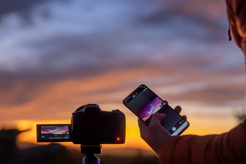 A person uses a smartphone to remotely adjust the settings on a Canon camera, which has a sunset scene on its screen. 