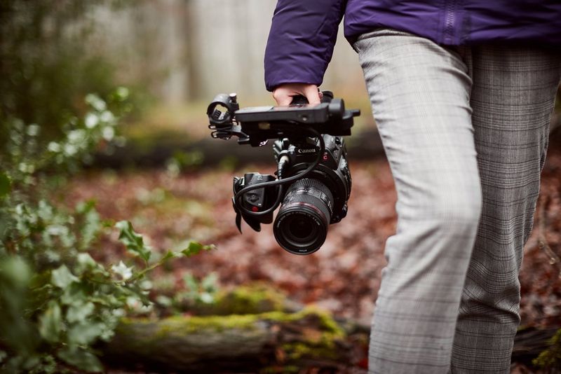 A woman walks through autumnal woodland, carrying a Canon EOS C200 video camera at her side.
