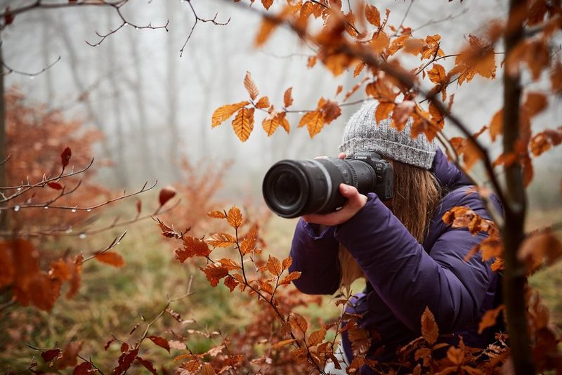 A young woman wearing a winter hat photographs autumnal leaves with a Canon camera and large lens. 