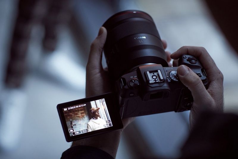 A pair of hands hold up a Canon EOS RP camera, with the vari-angle touchscreen folded out and up, showing a person standing in front of a window and brick wall.