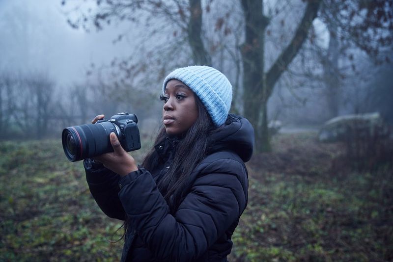 A person wearing a light blue winter hat and blue overcoat holds a Canon EOS RP in a misty woodland setting.