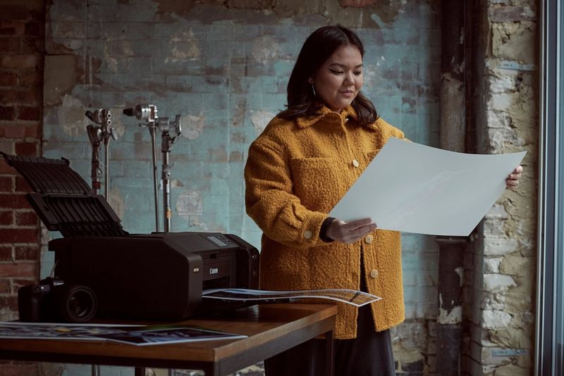 A photography student wearing a yellow coat studies an A3 image printed on a Canon imagePROGRAF PRO-300 printer, which is on the table next to her. 