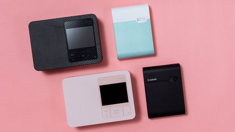 Black and pink SELPHY CP1500 printers and green and black SELPHY Square QX10 printers on a pink flat surface.