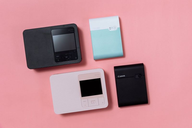 Two Canon SELPHY CP1500 printers sit next to two Canon SELPHY Square QX10 printers on a bright pink background.