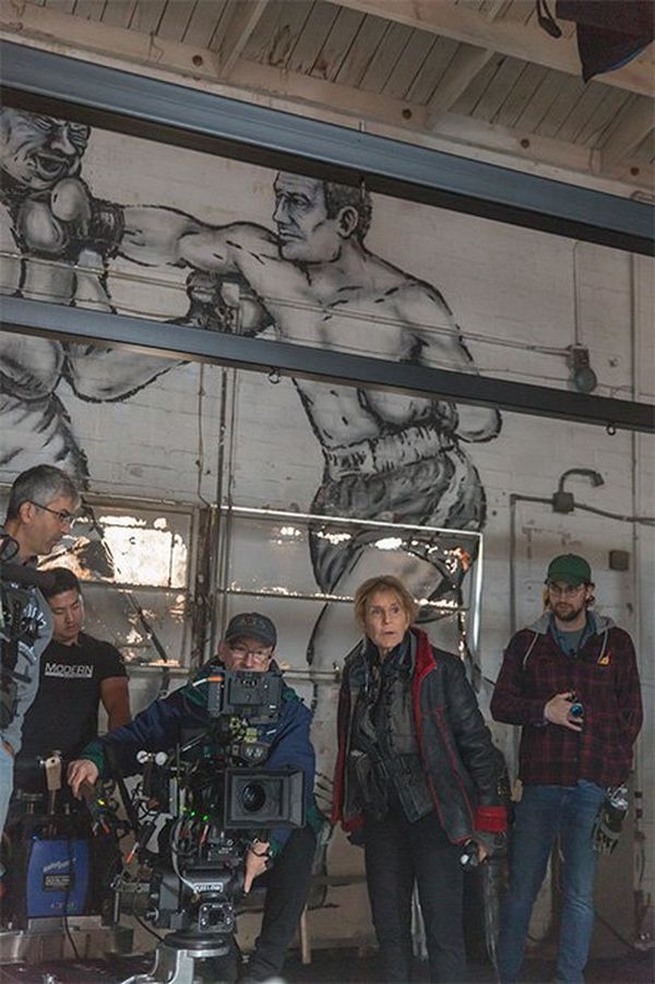 The camera crew at work in front of a large mural of a boxer landing a right cross on an opponent. Director Nigel Dick is seated behind a Canon EOS C700 FF, and cinematographer Nancy Schreiber stands to his left.