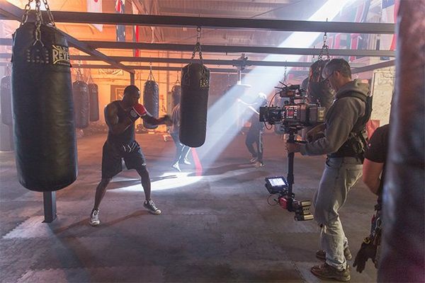A camera operator with a Canon EOS C700 FF on a Steadicam films a boxer using one of several punching bags in a boxing gym.