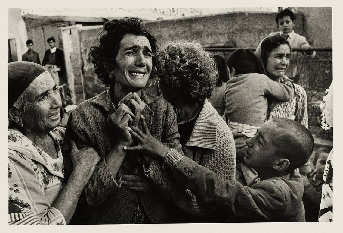 A grieving Turkish woman is comforted by others and clutches her hands to her chest as she mourns her husband, a victim of the Cyprus Civil War. In the background another woman can be seen weeping.